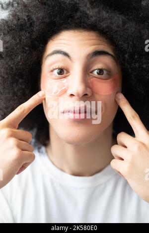 Cura del viso dell'uomo. Idratante per la pelle. Prodotto cosmetico anti-invecchiamento. Sorpreso bel ragazzo con lunghi capelli ricci marrone mostrando rosa hydrogel occhio patch o Foto Stock