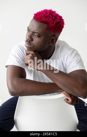 Uomo perplesso. Le vite nere contano. Progetti pensieri. Strategia mentale. Elegante serio ragazzo africano con capelli rosa brillante bianco t-shirt mano sul mento seduta Foto Stock