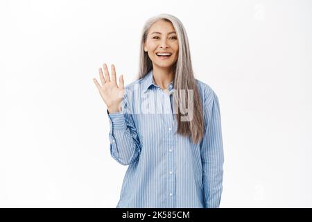 Donna anziana sorridente con capelli grigi, donna asiatica di mezza età che dice ciao, mano ondulata e saluto, in piedi su sfondo bianco Foto Stock