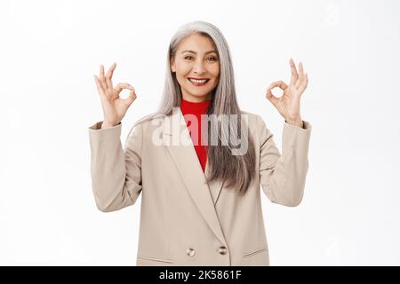 Molto bene. Donna d'affari asiatica soddisfatta mostrando bene, ok grande gesto, cenno di approvazione, sorridente compiaciuto, in piedi su sfondo bianco Foto Stock