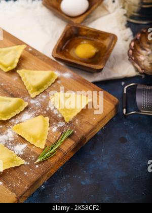 Primo piano. Ravioli crudi, utensili da cucina e ingredienti su un tagliere di legno su sfondo blu scuro. Ricette della cucina italiana, il processo di m Foto Stock