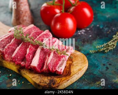 Il processo di cottura delle bistecche di carne. Pezzi crudi di carne selezionata - maiale, manzo, vitello su un tagliere di legno, spezie e pomodori freschi maturi su un buio Foto Stock