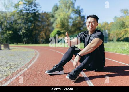 Ritratto di uno sportivo asiatico seduto su un tapis roulant dello stadio in un parco pubblico in una giornata di sole d'estate, l'uomo sta guardando la macchina fotografica e sorridendo dando pollici in su, corridore in abbigliamento sportivo all'aperto. Foto Stock