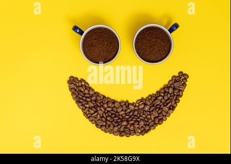Emoticon sorridente, labbra in chicchi di caffè e occhi in tazze di caffè, composizione su sfondo giallo. Foto Stock