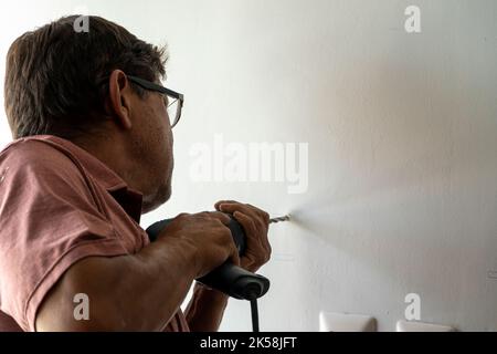 uomo latino nel suo 50s, perforando un muro con un trapano, muro bianco Foto Stock