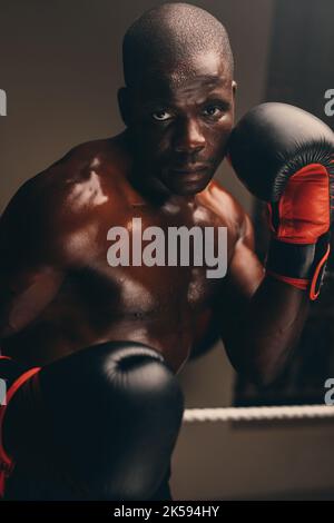 Pugile giovane e robusto che guarda la fotocamera mentre si trova in posizione di combattimento con i guanti da boxe. Determinato giovane uomo che pratica la sua tecnica di punzonatura Foto Stock