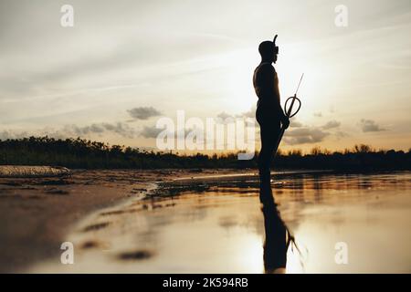 Vista laterale del pescatore di lancia che va pesca di spearfishing al tramonto. Silhouette ripresa da un subacqueo avventuroso in piedi in acqua di mare con un fucile in mano. Foto Stock