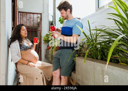 giovane moglie latina brasiliana incinta a casa seduto a bere caffè in una tazza rossa e dice al marito come si sente, mentre il marito è in piedi Foto Stock