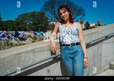 Una bella giovane donna dalla testa rossa che appoggia comodamente contro una parete bassa all'aperto in una giornata di sole. Aereo americano, CopySpace. Foto Stock