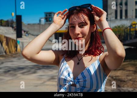 Un ritratto di bella giovane donna a testa rossa che regola gli occhiali da sole sulla sua testa. Spazio di copia. Orientamento orizzontale. Foto Stock