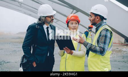 Ingegneri la donna asiatica e l'uomo arabo stanno parlando con il project manager in piedi presso il cantiere di costruzione insieme guardando intorno e controllando i documenti Foto Stock
