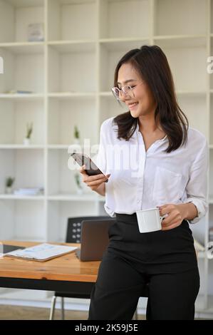 Ritratto, bella e sicura donna d'affari asiatica millennial o donna manager in piedi nel suo ufficio, utilizzando il suo smartphone per inviare messaggi mentre Foto Stock