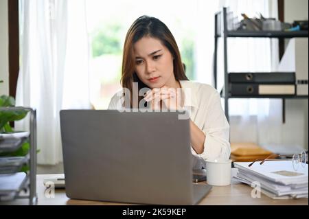 Pensieroso asiatico businesswoman si sta concentrando sulla pianificazione del suo nuovo progetto, guardando lo schermo del suo laptop, lavorando alla sua scrivania in ufficio. Pensivel Foto Stock