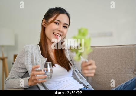 Giovane donna asiatica dall'aspetto bello seduta sul divano in soggiorno, con un bicchiere d'acqua e una foglia di lattuga verde. Concetto di stile di vita sano Foto Stock