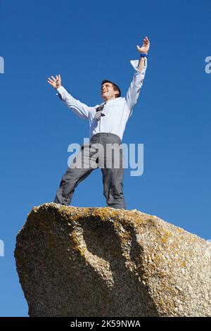 Raggiungere i pinnacoli del successo. Un giovane uomo d'affari in piedi con le braccia alzate in vittoria in cima a una scogliera. Foto Stock