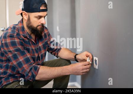 elettricista caucasico che tiene il cacciavite lavorando sulla spina elettrica su un impianto elettrico residenziale che installa la presa di alimentazione sulla parete grigia a casa Foto Stock