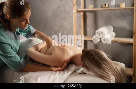 Massaggiatrice professionale o terapista manuale che massaggia la spalla della giovane donna, alleviando il dolore. Paziente femmina che ottiene il massaggio di corpo di rimedio in fisioterapia Foto Stock