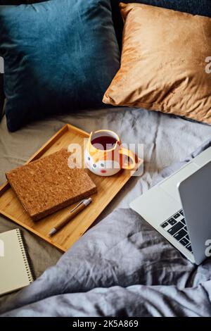 Casa accogliente, autunno caldo umore. Computer portatile, notebook e tazza di tè al vassoio su coperta nel letto all'interno della camera da letto. Accogliente autunno o inverno concetto. Foto Stock