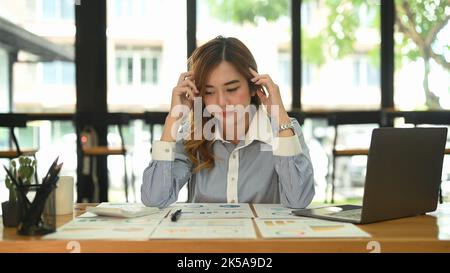 Una lavoratrice frustrata che tiene la testa e guarda documenti finanziari, pensando di trovare la soluzione al problema del lavoro Foto Stock