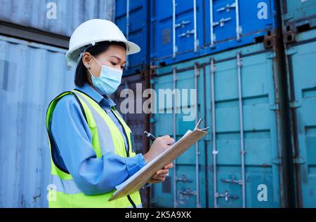 Covid, la logistica di spedizione e la scrittura del dipendente durante l'ispezione in un magazzino di contenitori. Donna di ingegnere industriale asiatica con note di ecommerce e. Foto Stock