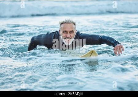 Uomo anziano, acqua e sport surfista su una spiaggia con sorriso formazione per fitness e salute o hobby all'aperto. Anziano maschio pagaiando in tavola da surf Foto Stock