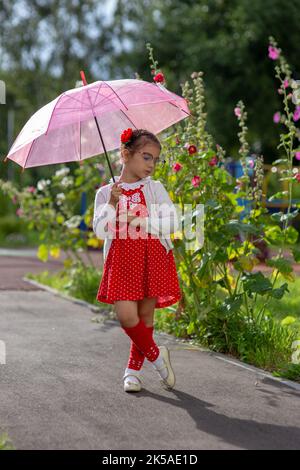 Una bella bambina in abito rosso e blusa bianca sotto un ombrello rosa si alza in un giorno d'estate. Foto Stock