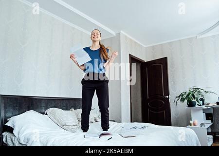 Eccitato giovane donna allegra ha ricevuto una lettera, è assunta, saltando sul divano a casa, ottenendo le buone notizie, ritenendo la gioia, ballando, ridendo, fare Foto Stock