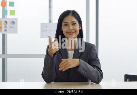 Giovane dipendente asiatico femminile in tuta grigia sedersi rilassarsi con il viso sorridente, sollevare il pollice dito di fronte mentre si siede alla sua scrivania. C'era un documento di note e. Foto Stock