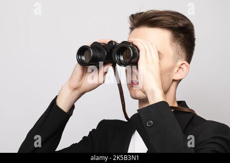 Ritratto di elegante giovane uomo che indossa tuta nera guardando attraverso binocoli. Studio girato su sfondo grigio Foto Stock