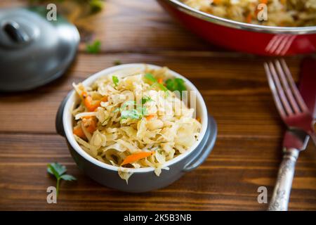 Cavolo brasato con carote, cipolle ed erbe in una ciotola di ceramica su un tavolo di legno. Foto Stock