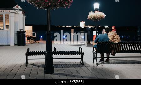 Una vista sul retro di una coppia seduta insieme su una panca vicino alla spiaggia di notte Foto Stock