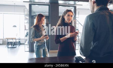 Una giornata di lavoro intensa presso un'accogliente caffetteria con clienti in piedi al banco che acquistano caffè e dipendenti cordiali che lavorano e parlano. Foto Stock