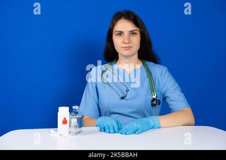 Un medico in uniforme blu con uno stetoscopio e guanti siede a un tavolo con farmaci. Foto Stock