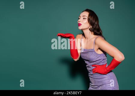 bruna giovane donna in guanti rossi e vestito viola sorridente mentre soffia aria bacio sul verde, immagine stock Foto Stock