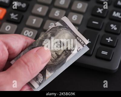 Un uomo che tiene in mano un conto di cento dollari con una calcolatrice sullo sfondo. Primo piano. Foto Stock