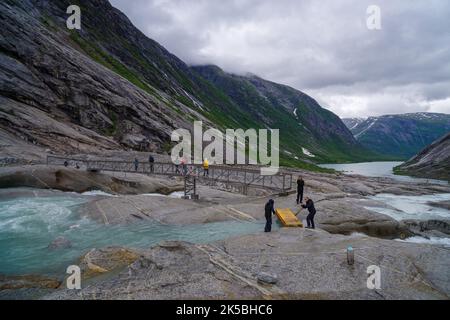 GJERDE, NORVEGIA - 3 LUGLIO 2022: Turisti che attraversano un ponte pedonale sulla loro strada per il ghiacciaio nigardsbreen sullo sfondo Foto Stock