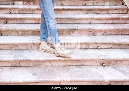 Gambe donna in jeans blu ritorto e stivali beige su scala in marmo rosa. Vista frontale Foto Stock