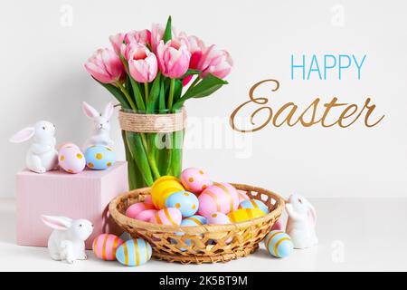 Concetto di biglietto d'auguri per le vacanze di Pasqua e di primavera. Tulipani rosa, coniglietti pasquali con uova colorate nel cestino. Uova di Pasqua dipinte in nido e tulipani con Foto Stock