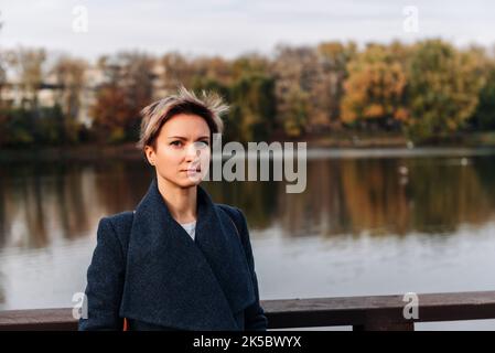 Una giovane donna in un pacco in autunno vicino al fiume guarda nella macchina fotografica Foto Stock