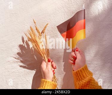 Happy Erntedankfest, Harvest Thanksgiving Day in lingua tedesca. Bandiera tedesca e mazzo di spighe di grano in mano su tessuto bianco beige. Giacitura piatta Foto Stock