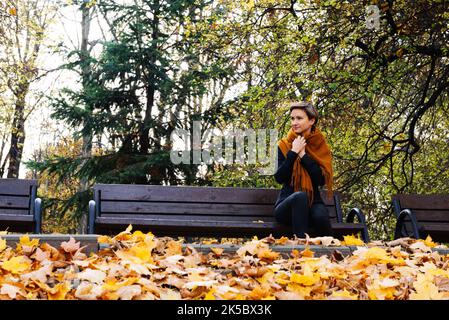 Una donna dai capelli corti si avvolge in una sciarpa seduta su una panchina in un parco autunnale Foto Stock