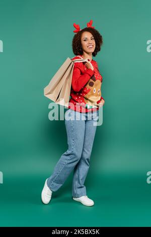 donna afro-americana sorridente in vestito di natale posando con le borse di shopping su sfondo verde, immagine di scorta Foto Stock