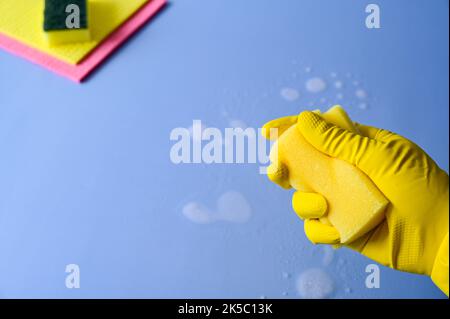 La mano con il guanto di gomma giallo tiene una spugna per la pulizia su sfondo blu. Foto Stock