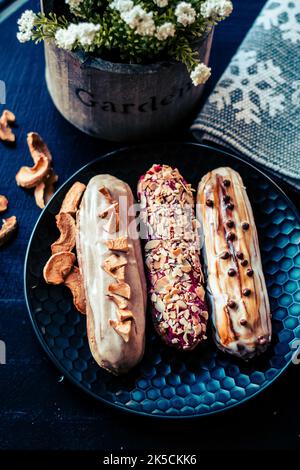 Vista dall'alto del tradizionale dessert francese con tre eclairs di mele secche, noci e caramello in una caffetteria su un tavolo da piatto scuro e fiori in una pentola Foto Stock