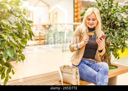 Ritratto di un giovane turista adolescente che visita la città e che trasporta le borse della spesa della carta mentre si appoggia alla vetrina di un negozio di moda, utilizzando il suo smartphone Foto Stock