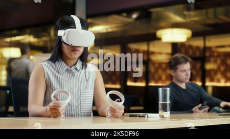 Donna asiatica che lavora in ufficio utilizzando visore VR e controller wireless, guardando i dati in realtà virtuale. Lavora in un ufficio moderno di un'azienda hi-tech con colleghi diversi. Tecnologia digitale nel cyberspazio Foto Stock