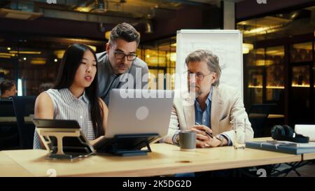 Team multietnico di lavoratori creativi che discutono di strategia aziendale o progetto di startup in un ufficio moderno utilizzando un computer portatile. Società di e-commerce. Lavorare sul progetto di avvio Foto Stock