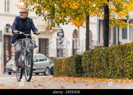Riprese all'aperto. Concetto di ciclismo. Foto integrale dell'uomo caucasico vestito in modo intelligente nel suo 60s con i capelli grigi in bicicletta da città nel parco dove le foglie stanno cambiando il loro colore in giallo. Foto di alta qualità Foto Stock