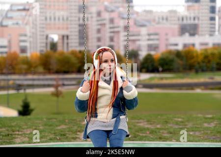 Giovane donna con lucchetti in cuffie wireless che oscillano sull'altalena, ascoltando musica nel parco cittadino. Bella femmina con capelli colorati multicolori Foto Stock