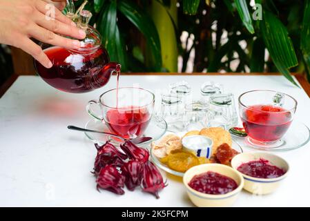 Mani che tengono teiera di vetro con tè di ibisco con tazze e frutta cristallizzata e marmellata su un tavolo Foto Stock